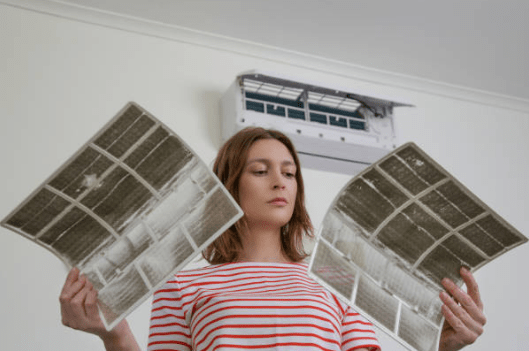A woman in a striped shirt holds up two extremely dusty mesh filters she has removed from the open indoor air conditioning unit mounted on the wall behind her.
