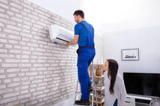 Standing on a ladder, a technician in blue work overalls aligns a white indoor mini-split unit on a light brick wall as a woman watches.