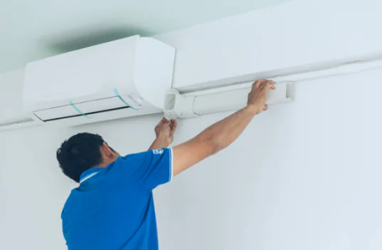 A technician in a blue shirt installs a white plastic conduit cover over the refrigerant line set connected to an indoor mini-split air conditioner.