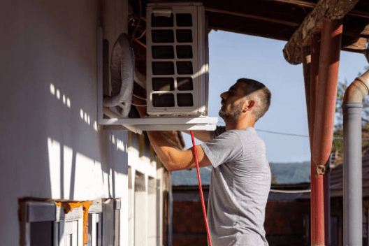 A technician in a grey t-shirt works to secure a white outdoor air conditioning condenser unit onto a wall-mounted metal bracket.