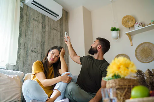 A man uses a remote to adjust a wall-mounted mini-split air conditioner while a woman sits on a sofa looking visibly frustrated by the heat.