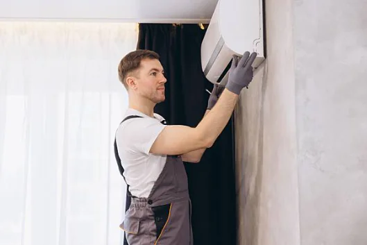 An HVAC technician wearing grey overalls and protective gloves carefully mounts a white indoor mini-split air conditioning unit onto a living room wall.