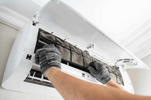 Gloved hands carefully slide a dust-clogged mesh filter out of the open service panel of a wall-mounted indoor air conditioning unit.