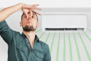 A man pinching his nose with a disgusted expression standing in front of a wall-mounted air conditioner emitting green wavy lines indicating bad odor.