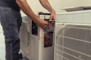 Close-up of a technician's hands working on the internal wiring and components of an outdoor air conditioning condenser unit.