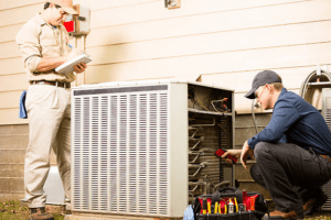 Two HVAC technicians servicing an outdoor air conditioning unit — one taking notes on a clipboard while the other examines the open unit's wiring and coils.